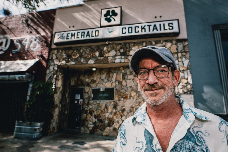 A medium-shot portrait of Mikey Brinkmann standing outside the Emerald Bar in St. Petersburg, Florida. Brinkmann is smiling at the camera, wearing a gray baseball cap, glasses, and a light-colored, short-sleeved button-down shirt featuring a blue octopus print. Behind them, the bar's entrance is visible, characterized by a distinctive, rough-hewn stone facade. Above the doorway, a horizontal sign with a shamrock icon reads 'EMERALD' on the left and 'COCKTAILS' on the right in black lettering.