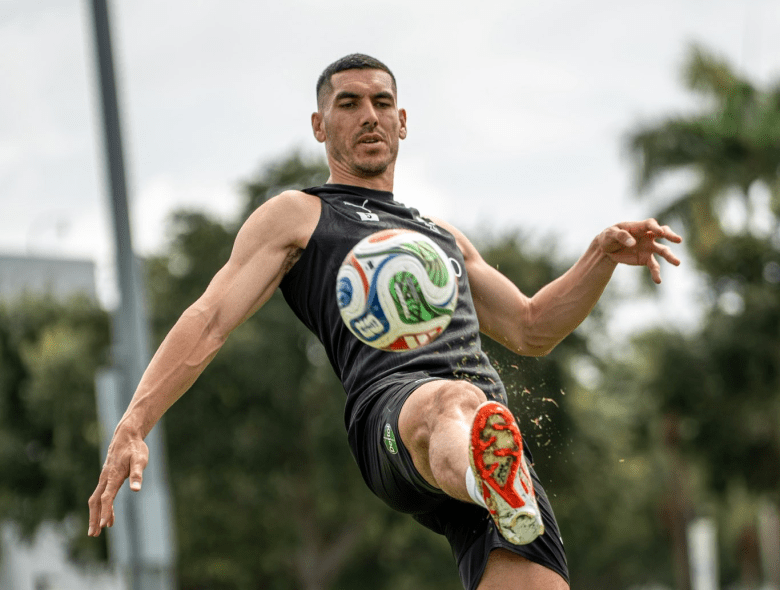 A medium-shot, eye-level photograph of New Zealand soccer defender Tim Payne captured during a training session. They have short dark hair and are wearing a black, sleeveless athletic training top. They are shown mid-air in a dynamic, athletic pose, striking a soccer ball with the instep of their right foot. The ball is positioned centrally in the frame, directly in front of their chest. The background is a soft, out-of-focus view of green trees and a pale, overcast sky, suggesting an outdoor sports complex.