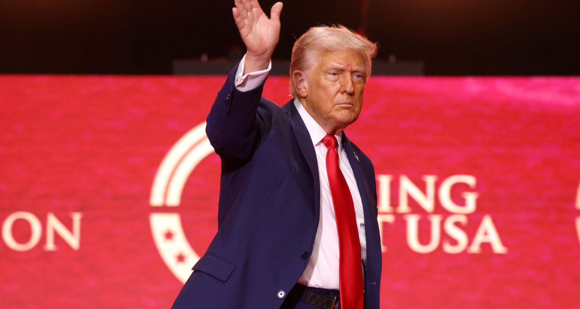 Donald Trump waving his right hand. He is wearing a red tie and standing in front of Turning Point USA red banner.