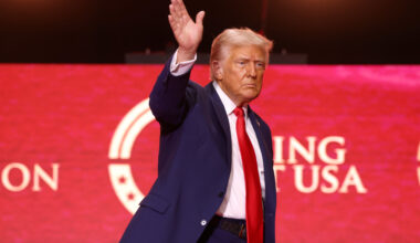 Donald Trump waving his right hand. He is wearing a red tie and standing in front of Turning Point USA red banner.