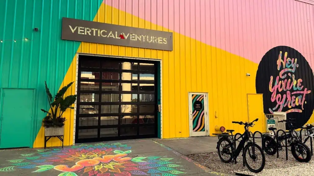 Photograph of a green, yellow, and pink building exterior featuring a large garage door with a "Vertical Ventures" sign above it. Several bicycles are parked outside, next to a colorful chalk mural on the sidewalk.