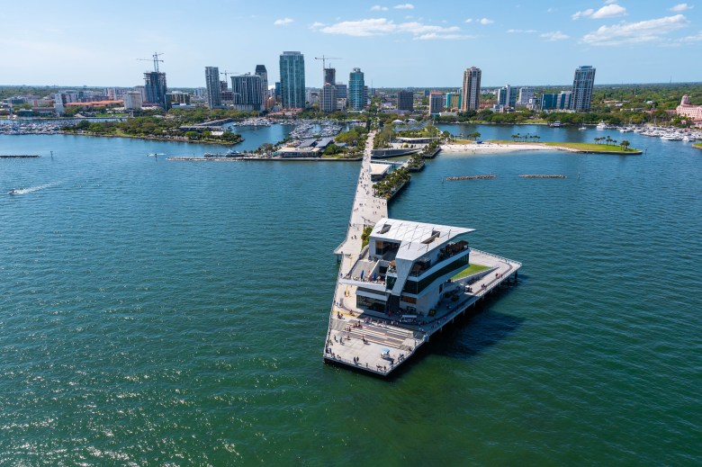 An aerial drone view of the modern St. Pete Pier jutting into the green waters of Tampa Bay. The iconic pointed pier head building sits at the end of the long concrete walkway, with the St. Petersburg city skyline and construction cranes visible in the background under a clear sky.