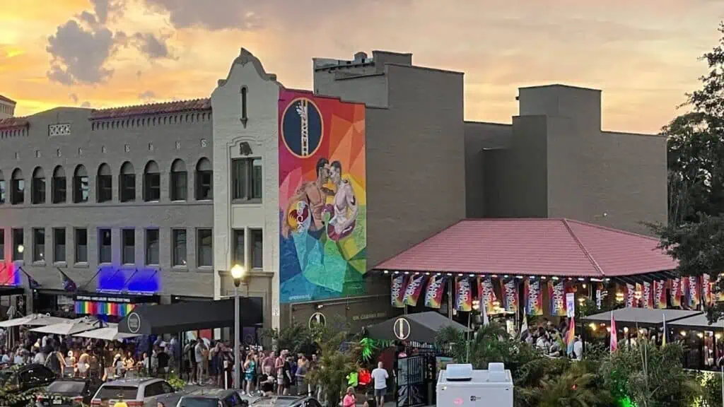 exterior of a hotel with a colorful mural on the far side. A line of people forms around the block to get in the front door. It's golden hour.
