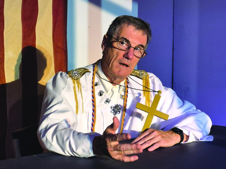 Dave Malloy in white judge's robes with ornate beading holds a cross necklace while seated in front of an American flag for Lab Theater Project's "The Cross & The Saber"