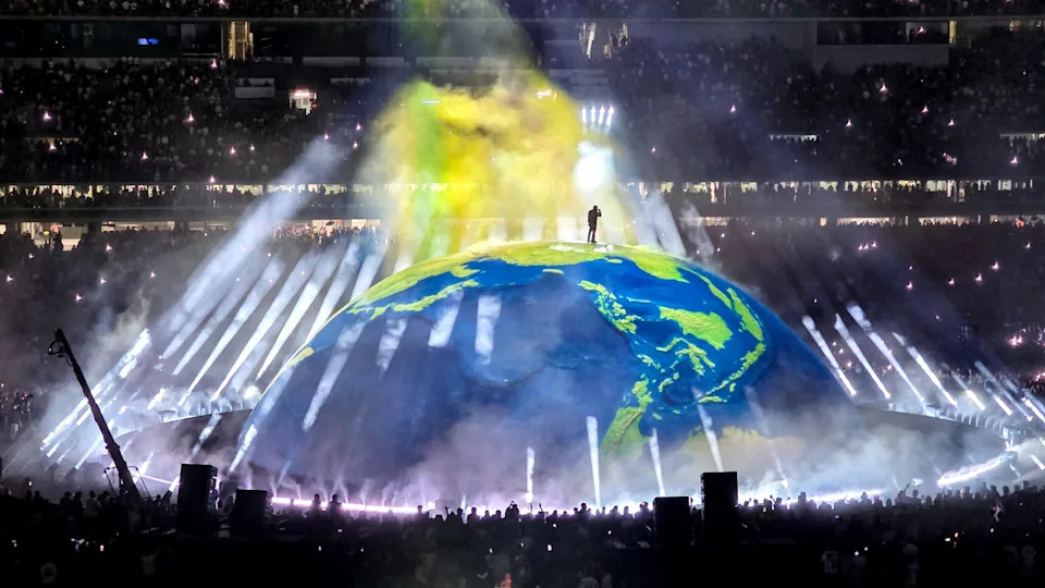 Ye stands on a large, illuminated globe surrounded by beams of light and smoke effects in a packed stadium.