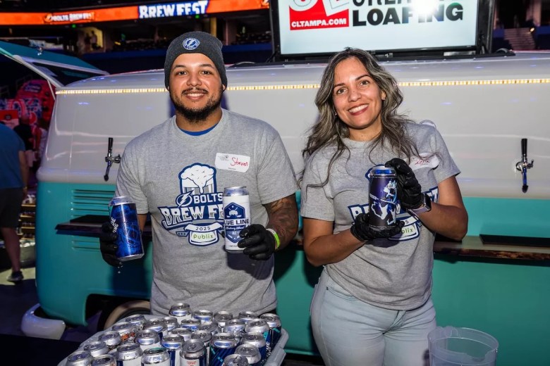 A medium-shot, indoor photograph of two volunteers smiling at the camera while working a booth at the Bolts Brew Fest at Amalie Arena. On the left, a person is wearing a gray 'Bolts Brew Fest 2025' t-shirt, a dark gray beanie, and a white name tag that reads 'Steve.' They are holding a blue aluminum beer can in their right hand. On the right, a person with long, wavy brown hair is wearing a matching gray event t-shirt, black gloves, and light-colored jeans. They are also holding a blue aluminum beer can. Behind them is a large, light-blue vintage-style van, which serves as the backdrop for their station, featuring a 'CLTAMPA.COM' logo sign on top.