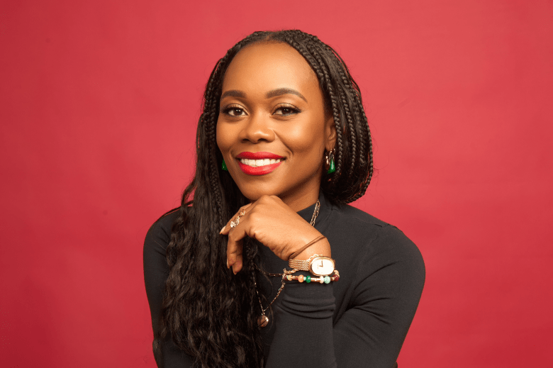 A medium-shot portrait of Tampa artist Melissa Koby. Koby is looking directly at the camera with a confident, welcoming smile. They have long, dark hair styled in braids, and are wearing a high-collared black long-sleeved top. Koby’s accessories include a delicate gold chain necklace, a watch on their left wrist, and a layered beaded bracelet. The background is a solid, warm-toned deep-red studio backdrop that makes the subject pop, creating a professional and artistic aesthetic.