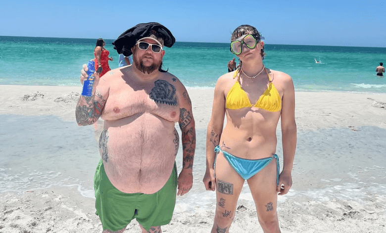 A medium-shot, outdoor photograph of two band members from Dogsmiles at the beach. On the left, a person with a dark beard and many arm tattoos is wearing a black head covering, white sunglasses, and green swim trunks; they are holding a blue aluminum beverage can in their right hand. On the right, a person with short brown hair is wearing yellow-tinted snorkeling goggles, a yellow bikini top, and light-blue bikini bottoms. They are standing on a white sand beach with the turquoise ocean and a clear sky in the background.