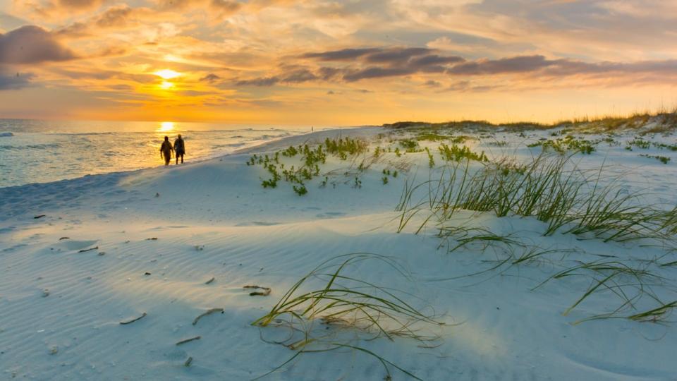 A wide shot of a sandy beach at sunset, with a golden sun setting over the ocean on the left. Two figures are silhouetted walking along the waterline. The foreground features white sand dunes with sparse green grass and low-lying plants. The sky is filled with orange and yellow clouds, reflecting the warm glow of the setting sun.