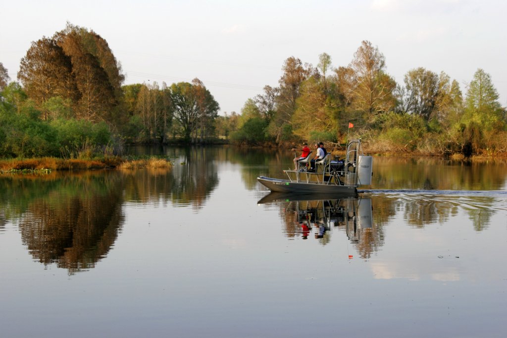 Three people riding an airboat on the Kissimmee River, with cypress trees reflected in the water.