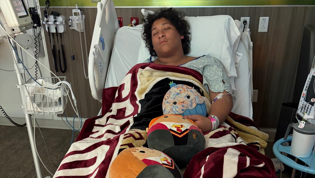 Joziah Thompson, a teenage boy, is seen in a hospital bed covered in blankets, with various medical equipment and stuffed animals around him.