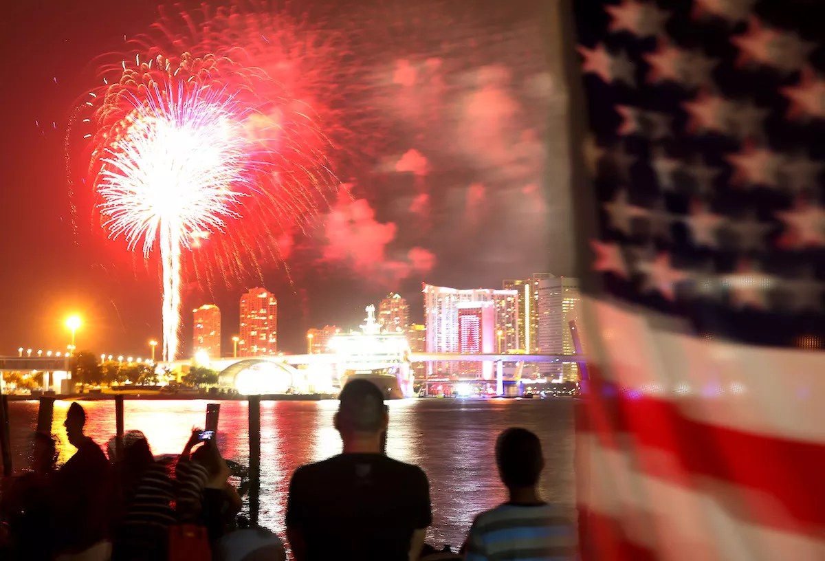 photo of silhouetted people watching a fireworks show over the water next to the Miami skyline. An American flag waves in the foreground to the right