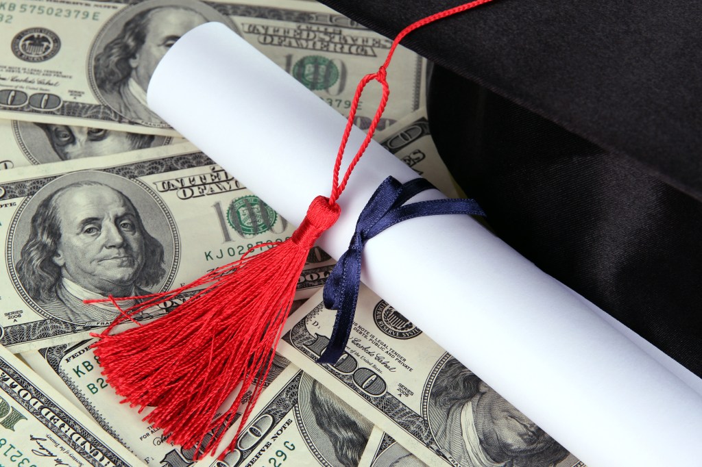 Graduation cap and diploma scroll with a red tassel and blue ribbon, lying on a bed of US hundred-dollar bills.