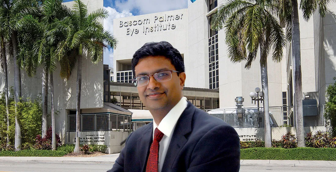 Dr. Ramulu in business attire and glasses stands in the foreground in front of the Bascom Palmer Eye Institute building, with palm trees and signage visible on the exterior in a sunny outdoor setting.