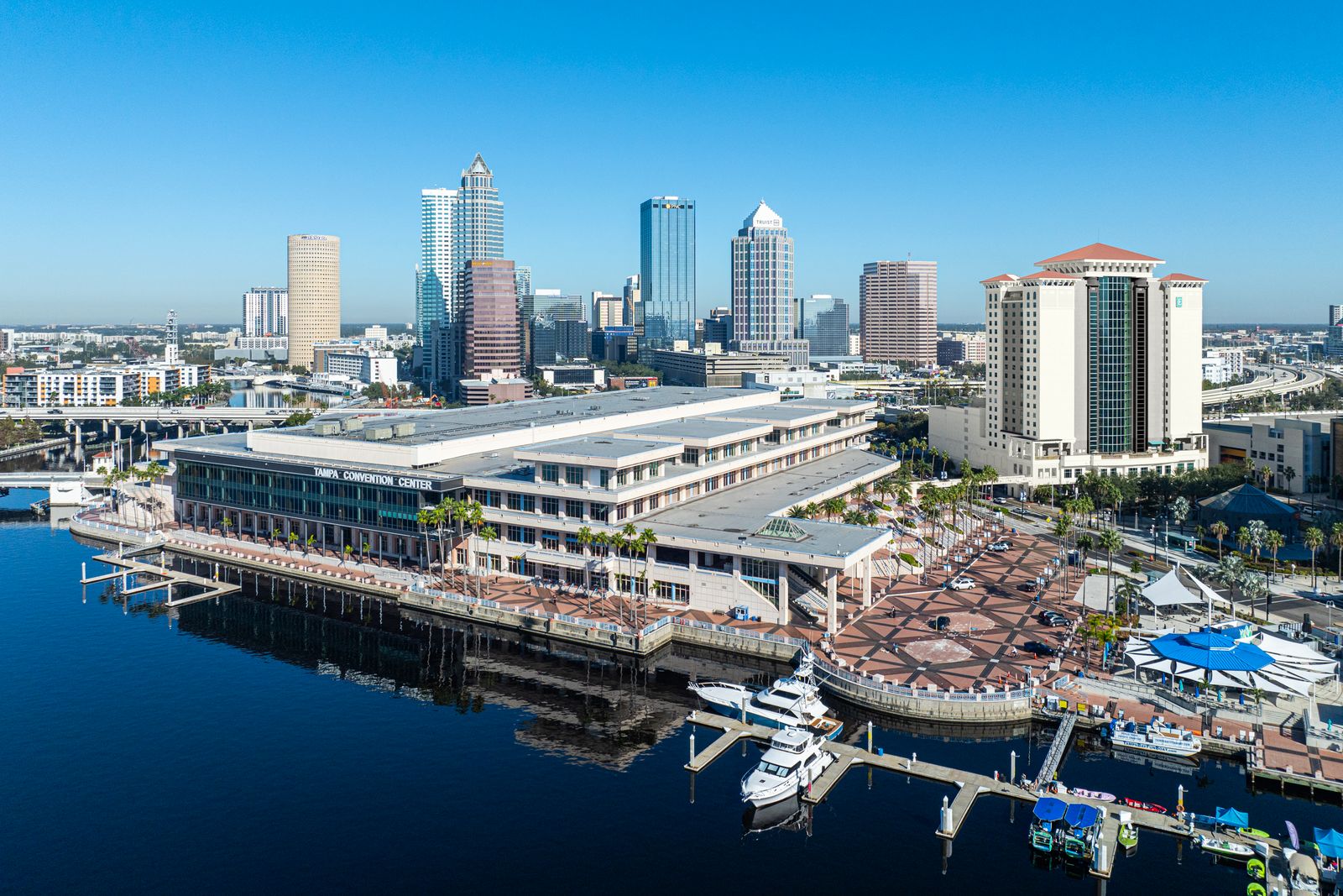 Aerial photo of the Tampa Convention Center on the waterfront with boats parked out front and modern glass exterior with Tampa city skyline in the background and blue skies in a walkable destination surrounding on the Tampa Riverwalk next to The Sail Plaza outdoor event space and bar.