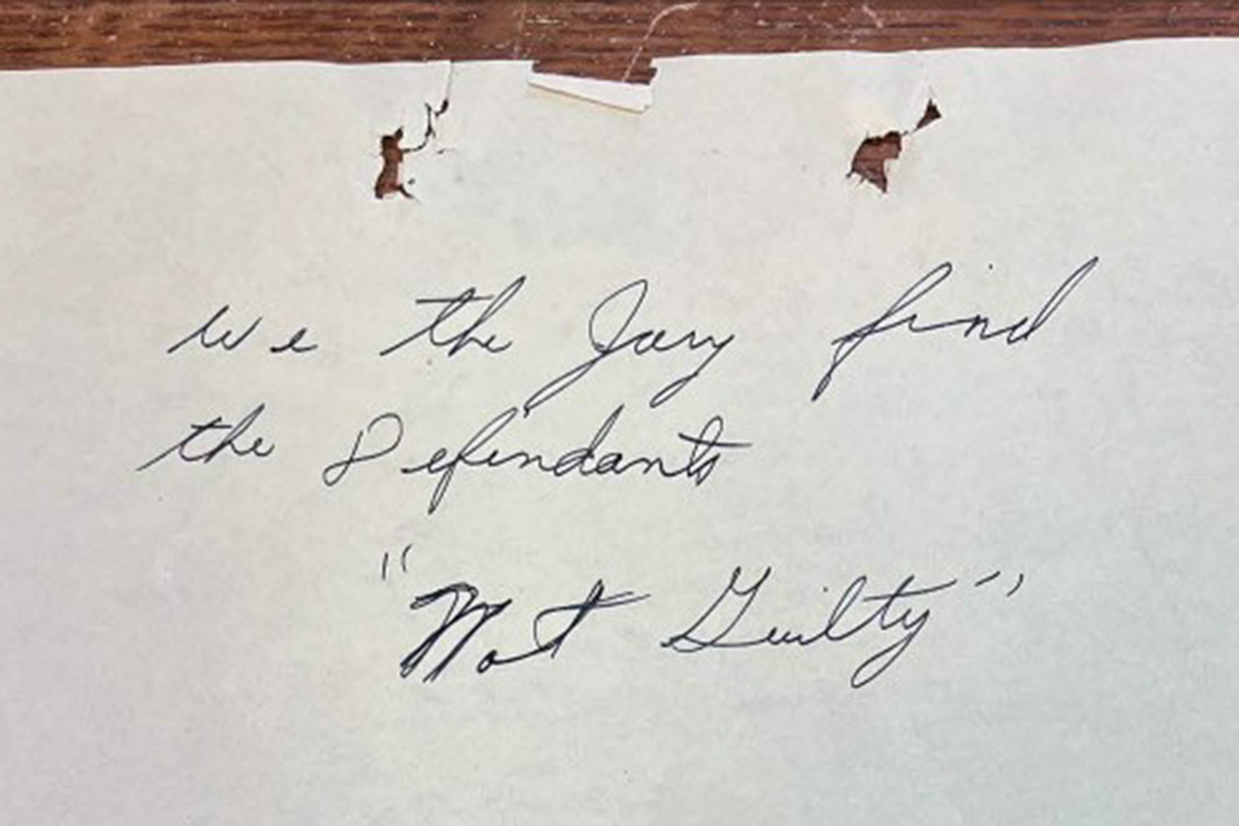 A close-up photograph of a piece of aged, slightly damaged paper featuring handwritten cursive text that reads, "We the Jury find the Defendants 'Not Guilty'".