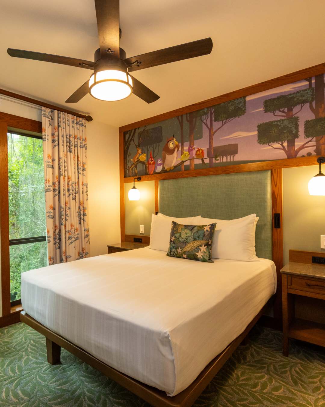 Another mural, this one a horizontal rectangle, shows the owl from Sleeping Beauty among other birds on a branch. The trees around them are pruned to be square-shaped. We see similar forest theming throughout the room, from the foliage-patterned carpet to the floral curtain pattern.