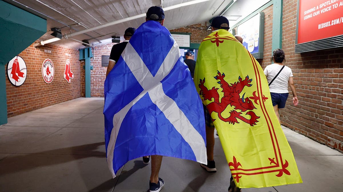 Two Scottish soccer fans walk through Fenway Park with flags draped over their backs.