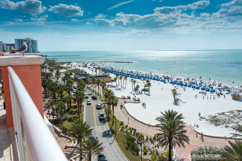High-angle aerial view of crowded Clearwater Beach in Florida with white sand, blue ocean, palm trees, and Pier 60 winding path.