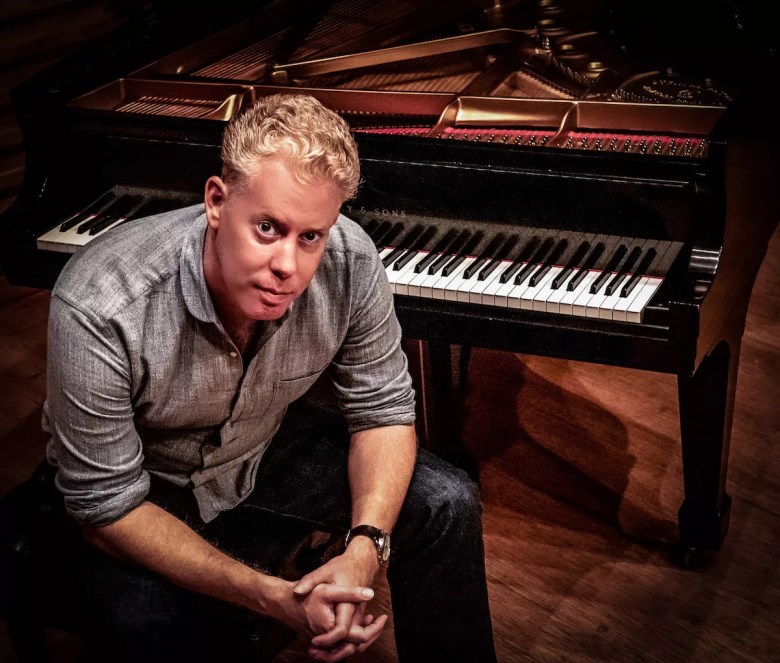 A portrait of composer and pianist Simon Lasky sitting in front of an open grand piano. The musician, with short blonde hair and wearing a grey button-up shirt with dark pants, sits with hands clasped, looking forward. The warm overhead lighting illuminates the piano keys and the polished wood interior of the instrument.