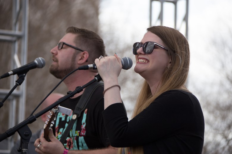 A medium-shot, live performance photograph of members of The Applebutter Express on stage at the Gasparilla Music Festival at Kiley Gardens in Tampa, Florida. In the foreground, a woman with long, straight light-brown hair is wearing black-rimmed sunglasses and a black long-sleeved shirt, singing passionately into a microphone. Beside them, to the left, a man with short brown hair, also wearing black-rimmed sunglasses and a black graphic t-shirt, is playing an acoustic guitar while singing into their own microphone. Both are illuminated by bright, soft outdoor light, with blurred trees and festival stage structures visible in the background.