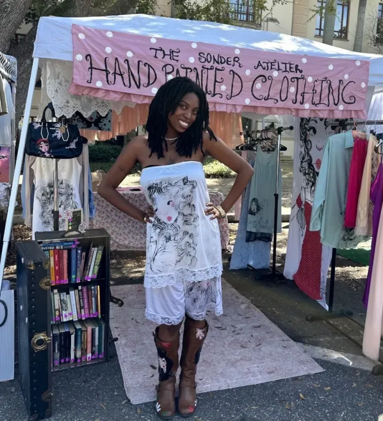 Jounte Armantrading poses outside a tent with the banner "The Sonder Atelier Hand Painted Clothing" wearing one of her designs, a frilly white tube top and shorts with a woman daintily painted on it