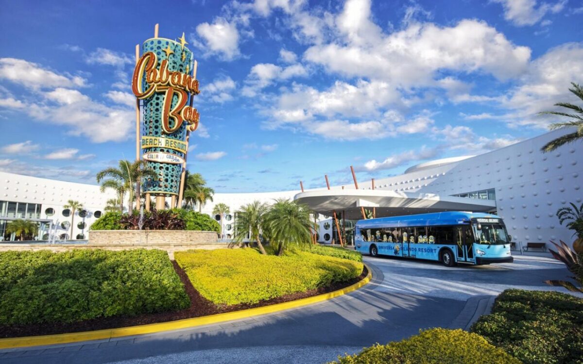 Cabana Bay Beach Resort entrance with a large colorful sign and an electric shuttle bus parked in front. The resort, part of Universal Orlando, features modern architecture and is surrounded by landscaped greenery under a blue sky.