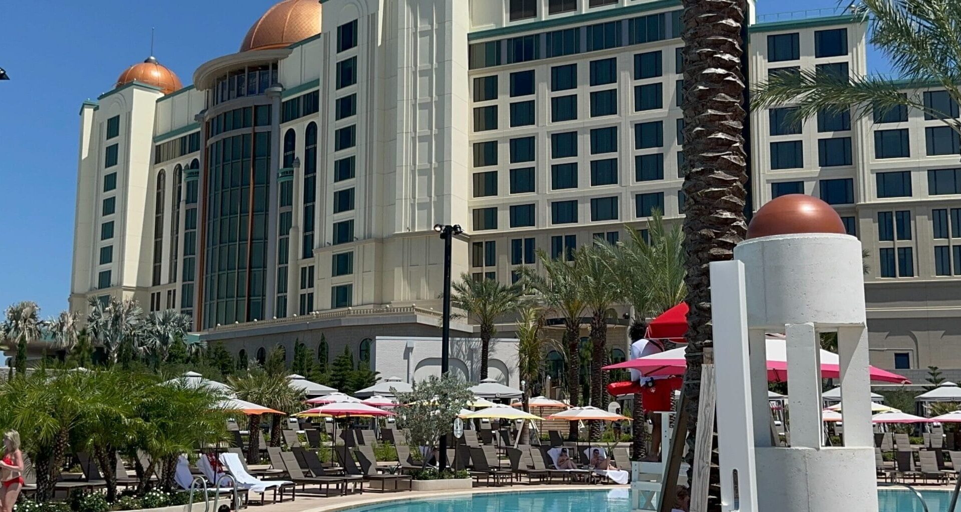 The Universal Helios Grand Hotel, with its domed rooftops, overlooks an outdoor swimming pool surrounded by lounge chairs, umbrellas, palm trees, and the clear blue sky—an epic universe of relaxation and luxury.