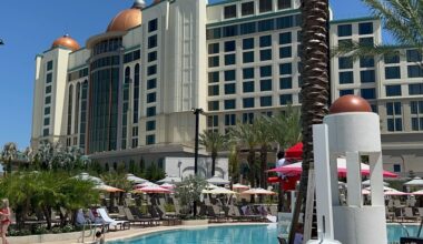 The Universal Helios Grand Hotel, with its domed rooftops, overlooks an outdoor swimming pool surrounded by lounge chairs, umbrellas, palm trees, and the clear blue sky—an epic universe of relaxation and luxury.