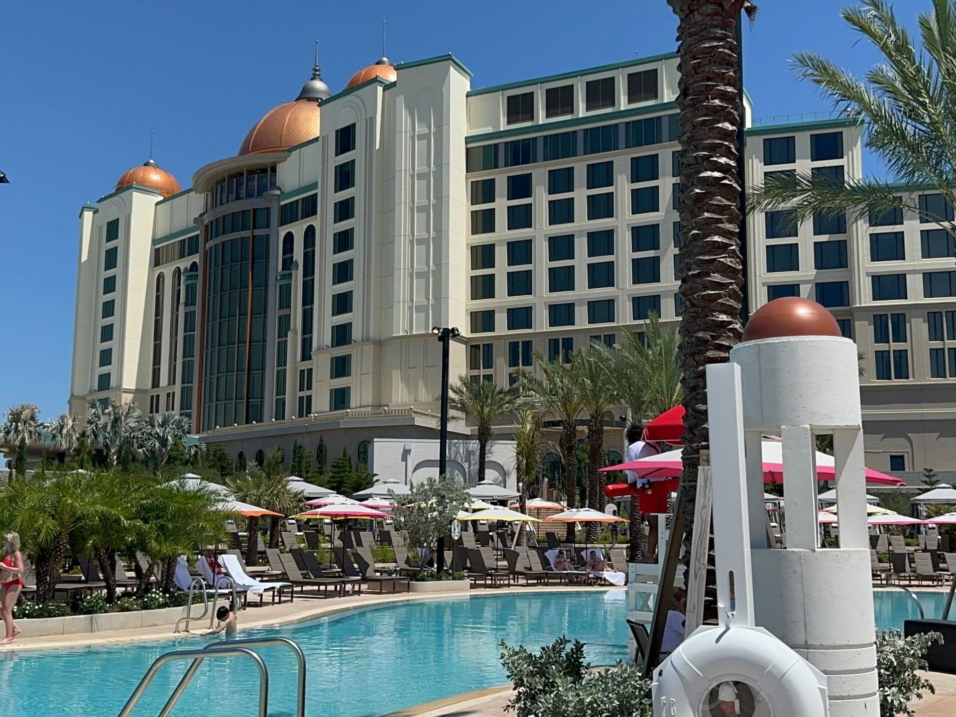 The Universal Helios Grand Hotel, with its domed rooftops, overlooks an outdoor swimming pool surrounded by lounge chairs, umbrellas, palm trees, and the clear blue sky—an epic universe of relaxation and luxury.