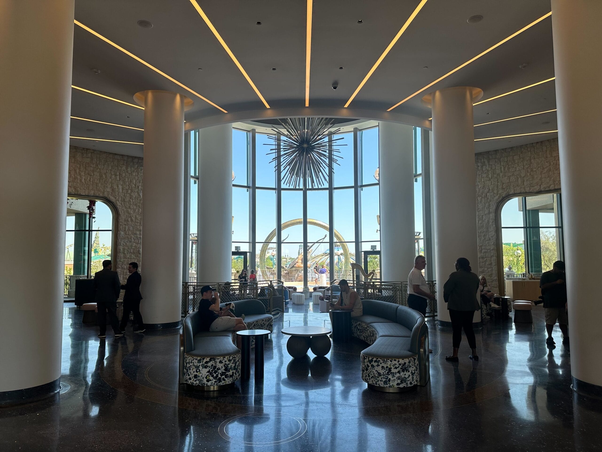 Modern hotel lobby at the Universal Helios Grand Hotel with tall windows, curved sofas, people relaxing, and a grand decorative light fixture—an inviting space inspired by the style of Epic Universe.