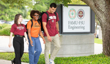 Three students walk together on a campus sidewalk beside a sign reading “FAMU-FSU Engineering.” The students wear university apparel and backpacks while talking beneath a large oak tree.