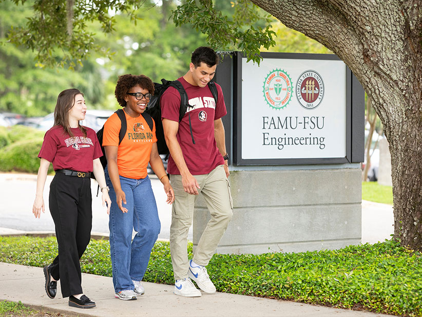Three students walk together on a campus sidewalk beside a sign reading “FAMU-FSU Engineering.” The students wear university apparel and backpacks while talking beneath a large oak tree.