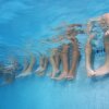 This photo, taken underwater, shows the lower legs of children who are sitting in a row along the edge of a swimming pool. Their legs, from the knees on down, are dangling in the pool's water.