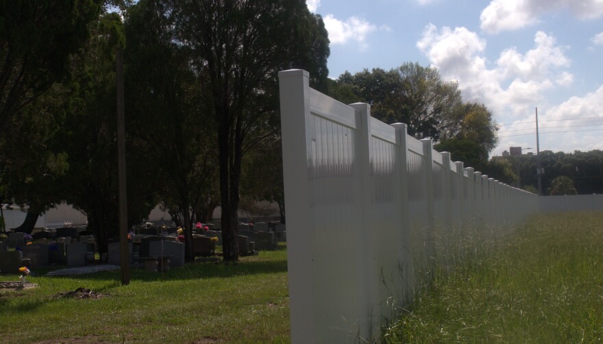 A tall white picket fence divides a lot of tall, unkempt grass from cemetery tombstones underneath a line of trees.