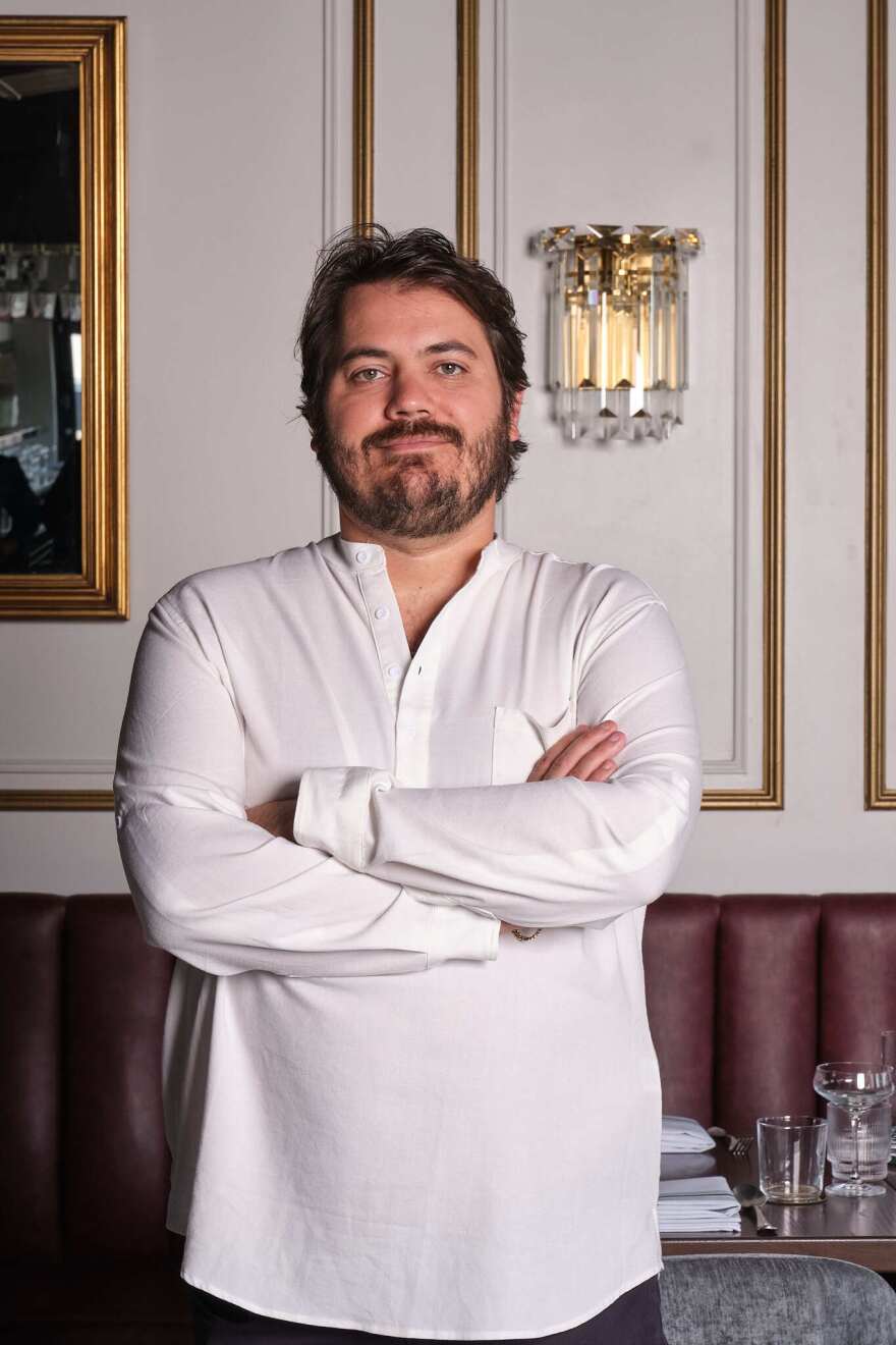 Man posing with his arms crossed while wearing a long-sleeve white shirt. He is at a restaurant, and his background consists of a white wall with details in gold, and a brown leather banquette seating.
