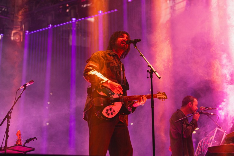 A vibrant live music photograph of Kevin Parker, frontman of psychedelic rock artist Tame Impala performing on stage at Benchmark International Arena in Tampa, Florida, on July 9, 2026. In the center, the musician is framed from the waist up, singing intensely into a microphone while playing a black and white Rickenbacker electric guitar. They have shoulder-length unkempt dark hair and a beard, and are wearing a dark overshirt layered over a lighter t-shirt and dark trousers. To the right, a second band member is partially visible playing a keyboard setup. The performance space is deeply atmospheric, saturated in bright pink, purple, and orange stage lighting, filled with thick theatrical smoke, and illuminated by glowing vertical light beams in the background.