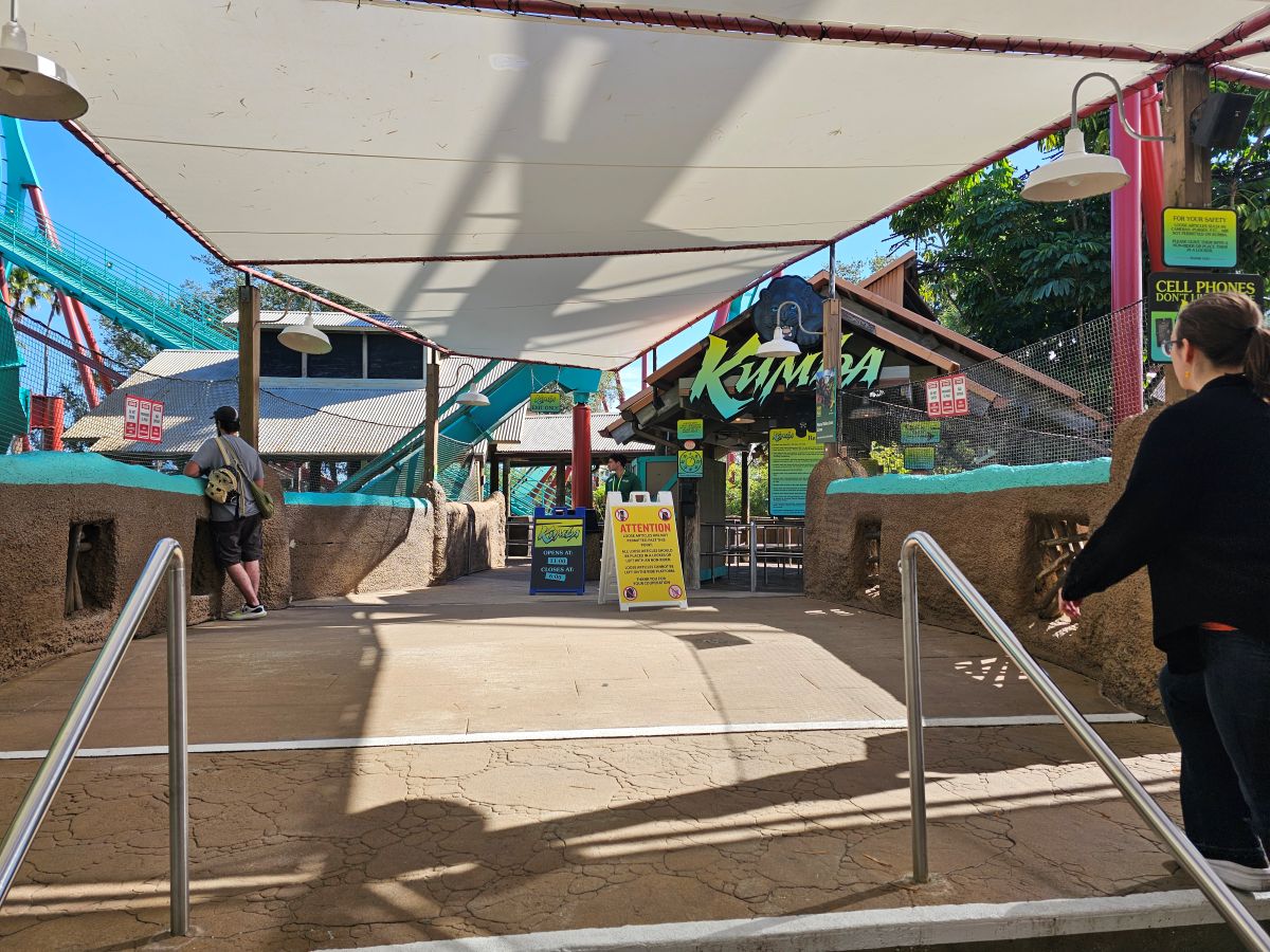 The paved entrance queue bridge to a roller coaster, showing the overhead shade canopy, entrance sign, and a yellow warning sign.