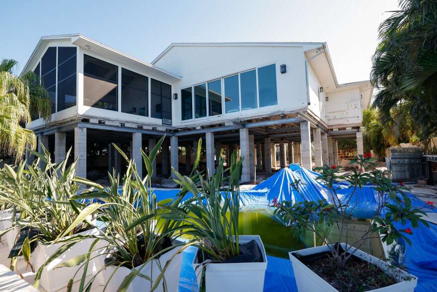 A house being lifted into the air by concrete pillars stationed along the foundation. In the foreground there are green plants in white planters and a blue tarp laid out over the ground.