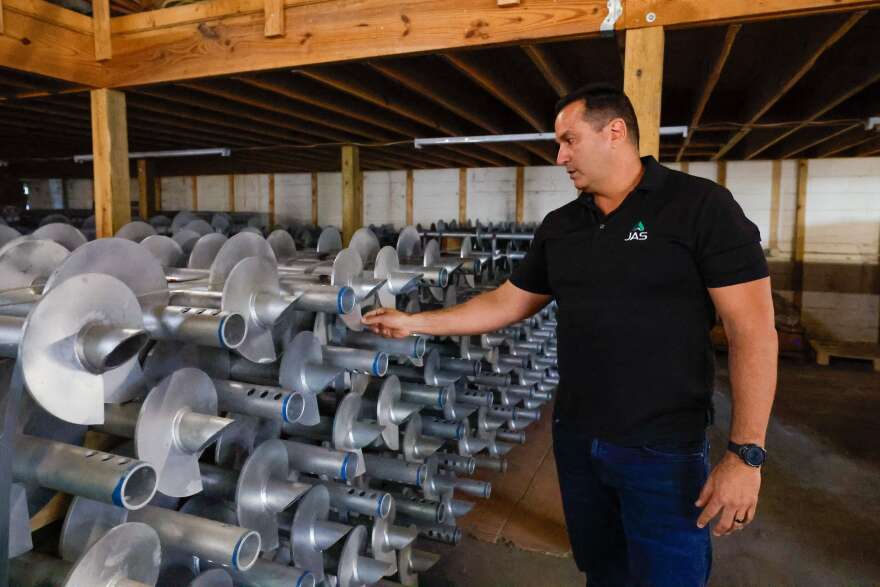 A man wearing a black polo shirt and blue jeans points to a stack of silver helical pilings, which look like long silver beams with a pointed end and spiraling metal components.