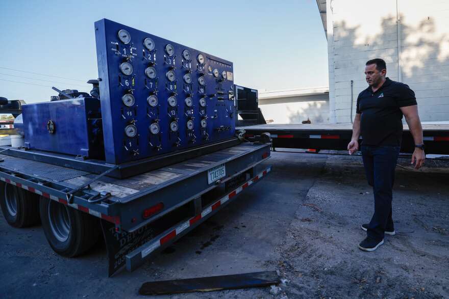 A man wearing a black polo shirt and blue jeans stands in front of a flatbed trailer with a blue piece of equipment strapped to it.