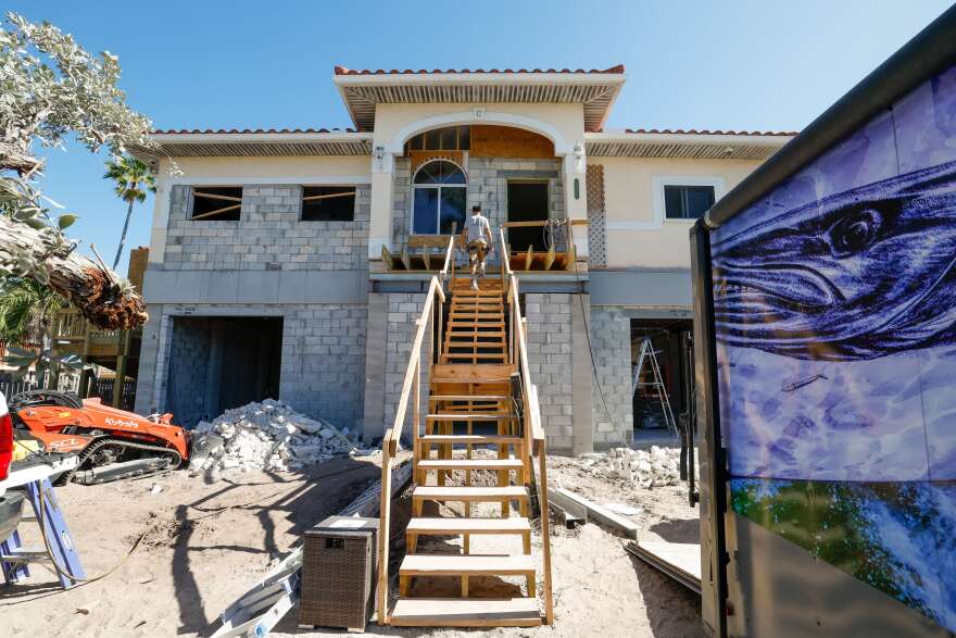 A house built up an entire story. The bottom level has exposed gray stone, and there’s a long wooden stairwell leading up to his front door. A man walks up the stairs and is close to the top.