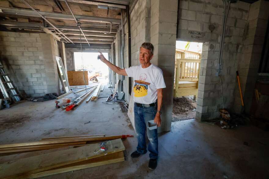 A man in a white t-shirt with a colorful design on it, and wearing blue jeans, stands in the bottom level of his house underneath where it’s been elevated. He points to the exposed gray stone. There are some construction materials and wooden beams lying on the floor.