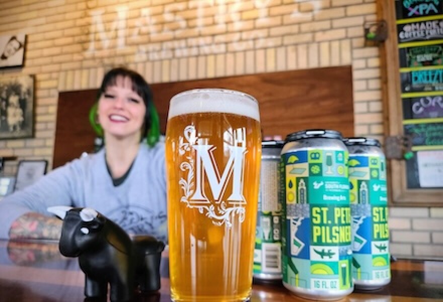 A bartender with green and brown hair folds their hands on a bar and is in the background slightly out of focus. In the foreground is a tall pint class with a M on it, filled with a lightish beer with a foam head. Next to it is a four-pack of tall boy-sized beer cans with the St. Pete Pilsner logo on it.