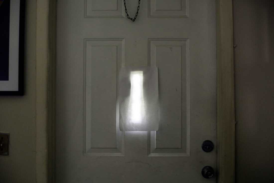A sheet of white paper covers a small narrow window in the middle of the white front door of Farah Larrieux's home, which is photographed from the inside.