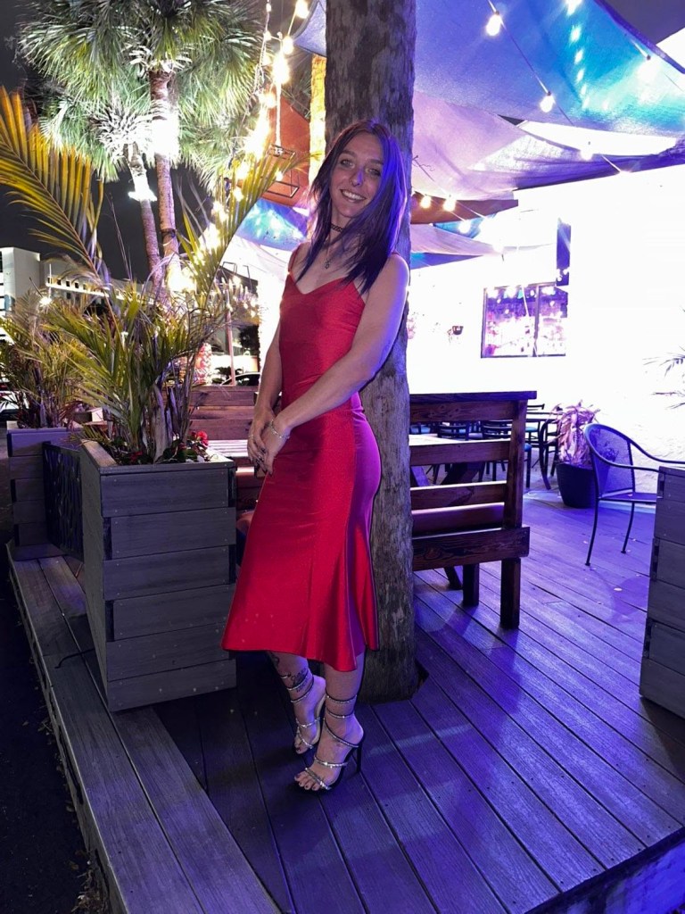 Brittany Clark in a red dress, leaning against a tree at a night outdoor venue.