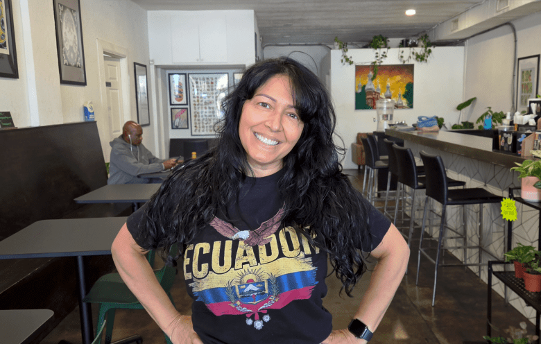 A portrait photo of Sandra Carson standing inside WeVegan Cafe in South Tampa, Florida, on June 25, 2026. Positioned center frame in a medium shot, Carson smiles warmly at the camera with hands resting on hips, long dark curly hair, and wearing a black t-shirt featuring a colorful graphic that reads "ECUADOR." The cafe interior features dark dining tables on the left, a counter with high bar stools and potted green plants on the right, and bright artwork decorating white walls in the background.