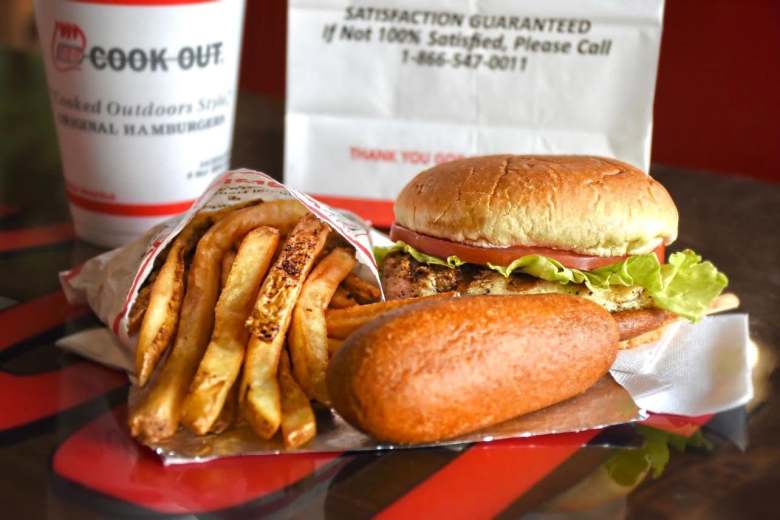 A Cook Out restaurant fast-food meal featuring a grilled chicken sandwich with lettuce and tomato, a side of french fries, a corn dog on foil, a branded drink cup, and a white paper takeout bag on a red table.