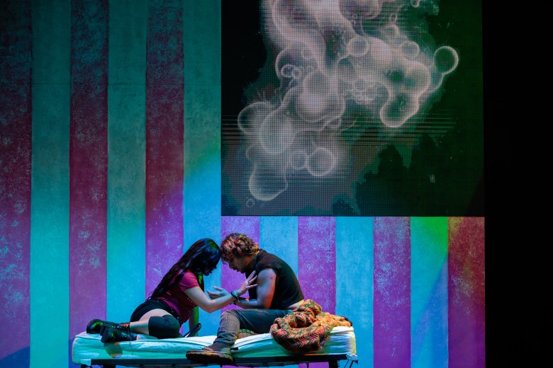 Two actors embrace in front of a dark American flag with smoke where the stars should be
