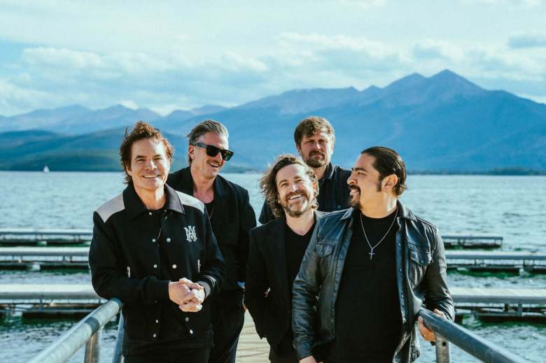 A promotional photo of the rock band Train, featuring lead singer Pat Monahan and his four bandmates smiling together on a wooden pier. They are dressed in casual black jackets and shirts. In the background, a wide, calm lake stretches toward a majestic range of blue mountains under a bright, partly cloudy sky.